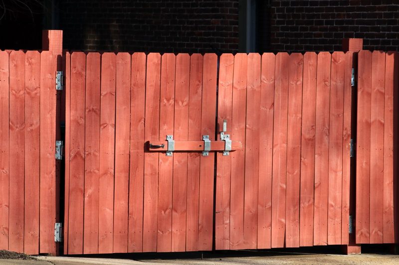 Red-stained wooden double gate with a galvanized drop latch and hinges, set in a board fence