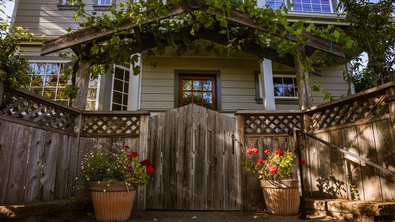 Weathered wooden gate with an arched top between lattice-topped fence panels and potted flowers, in front of a bungalow