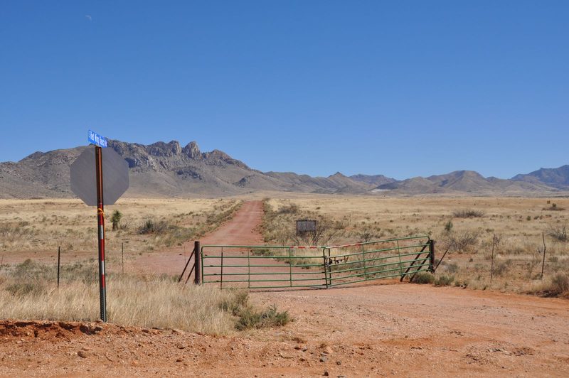Steel ranch gate closing a red dirt road beside a stop sign, with grassland and mountains behind