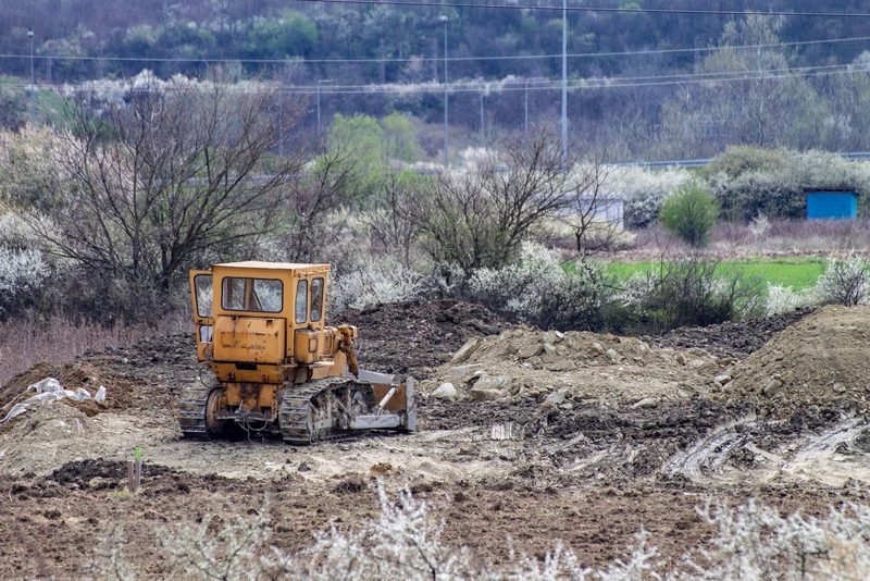 Crawler bulldozer on bare soil among brush and bushes on a rural site, with hills behind