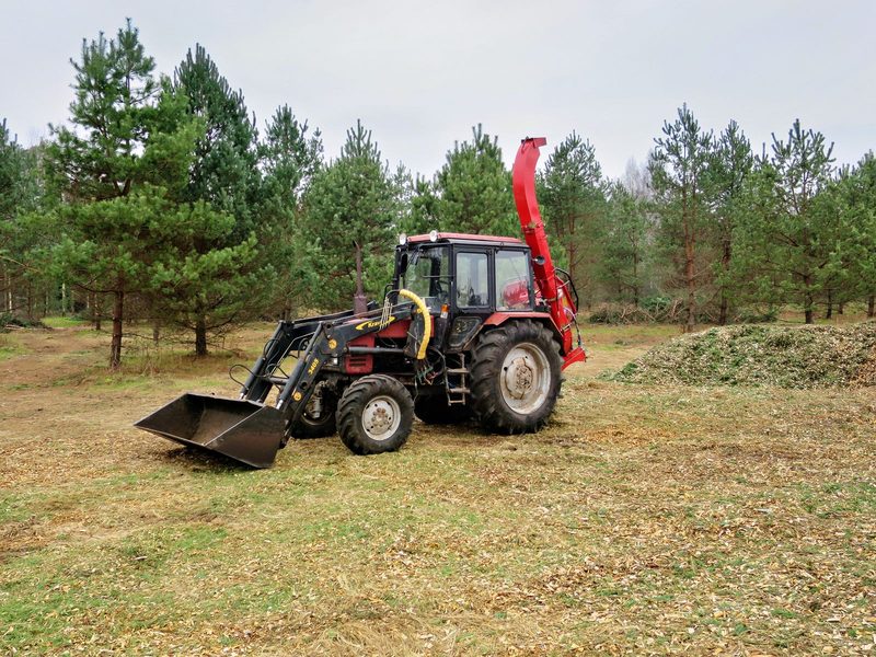 Red tractor with a front loader and a rear-mounted wood chipper in a pine clearing, piles of chips behind