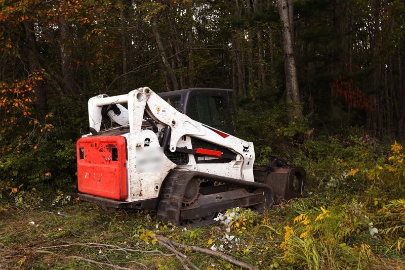 Compact track loader parked at the edge of cut brush in an autumn hardwood stand
