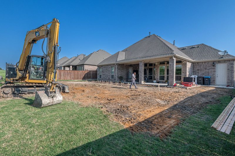 Excavator beside a freshly graded backyard behind a brick home, with form stakes set near the patio